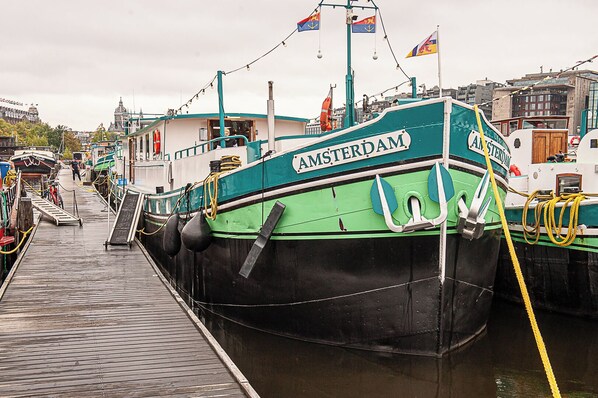 A docked boat named "AMSTERDAM" with a ramp leading to the dock, surrounded by other boats and buildings. - Amsterdam Hotelboat  (Amsterdam)