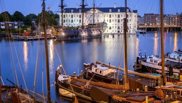 A harbor with several sailing ships docked, a prominent white building with a dark roof, and a calm water surface reflecting the structures.
