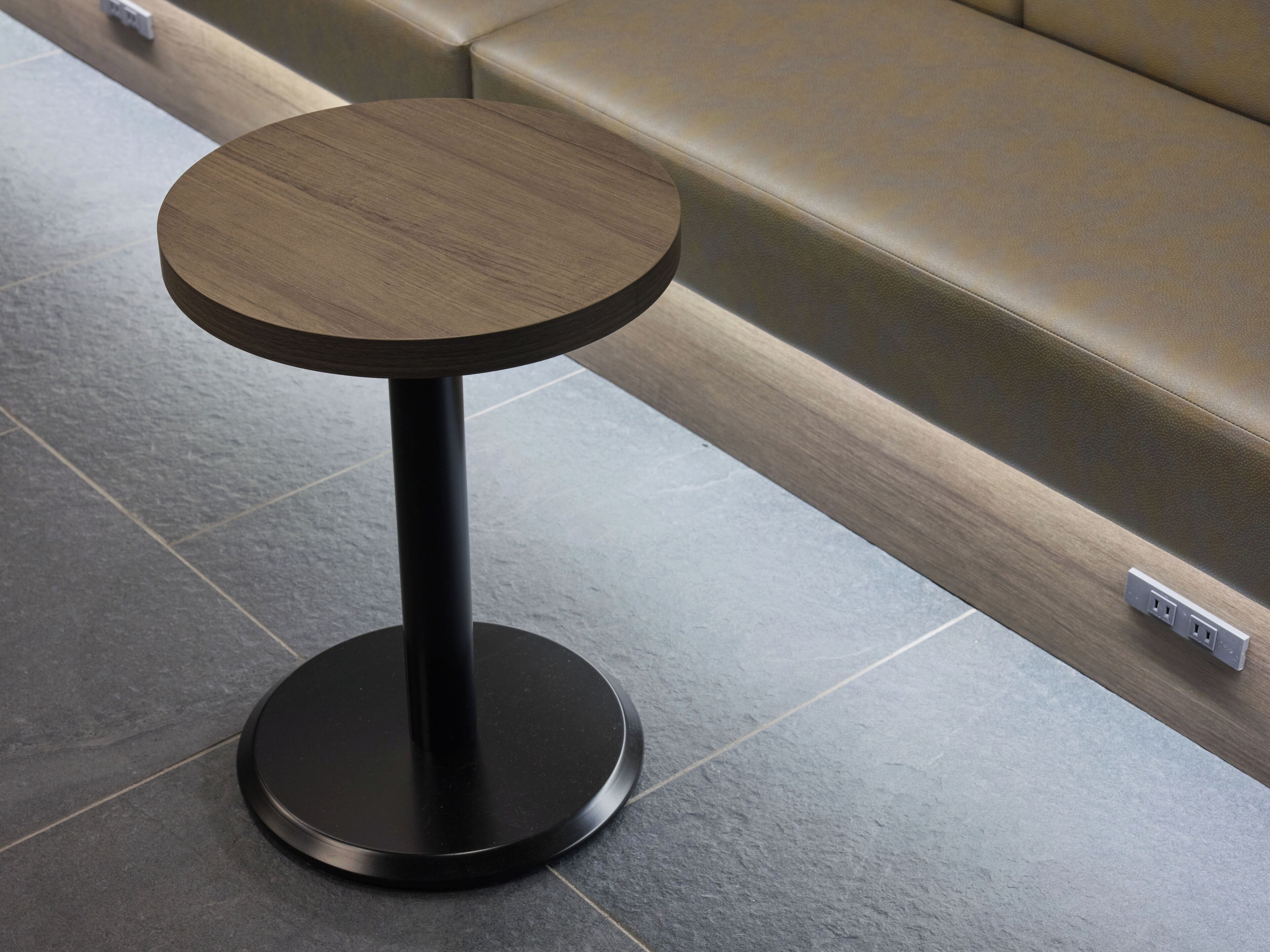 A round table with a dark top and a black base, positioned on a tiled floor next to a beige sofa.