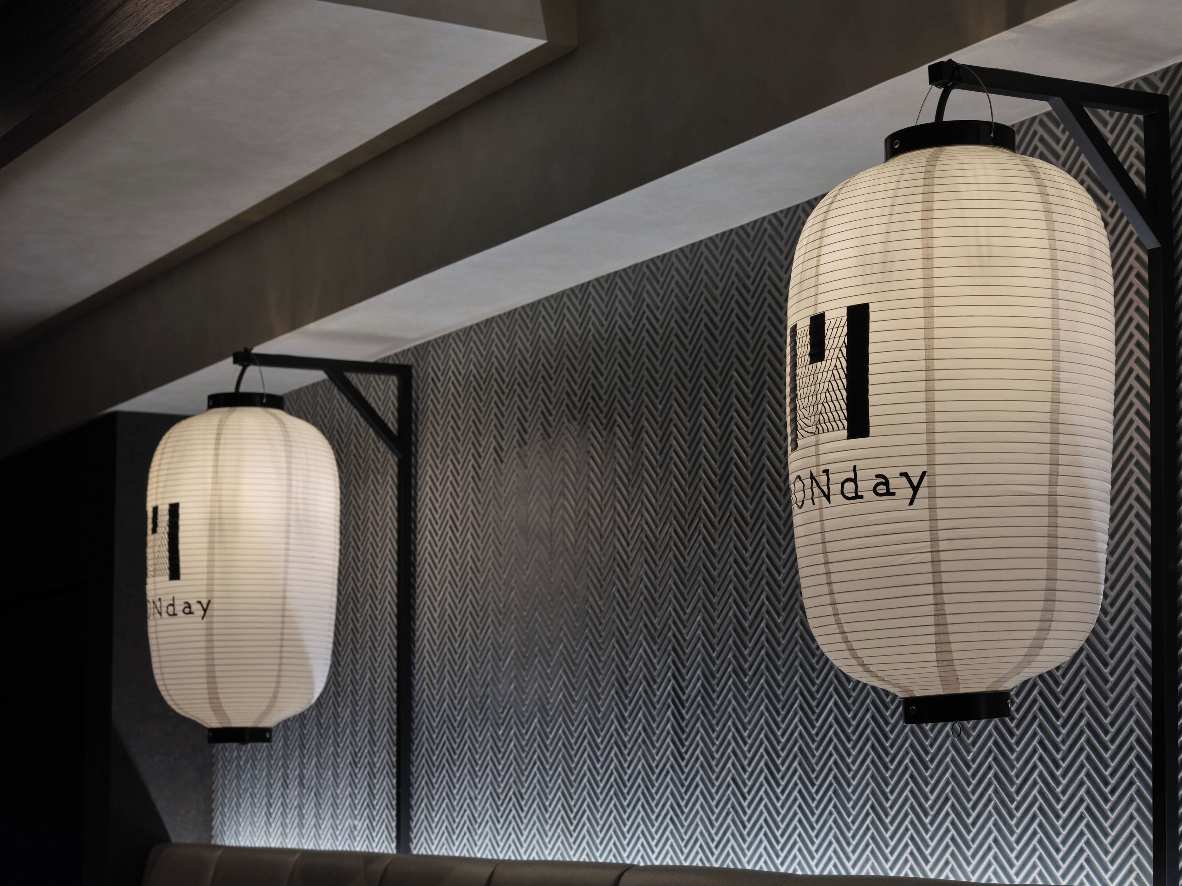 Two hanging paper lanterns with the word "Monday" on them, suspended from the ceiling in a room with a herringbone patterned wall.