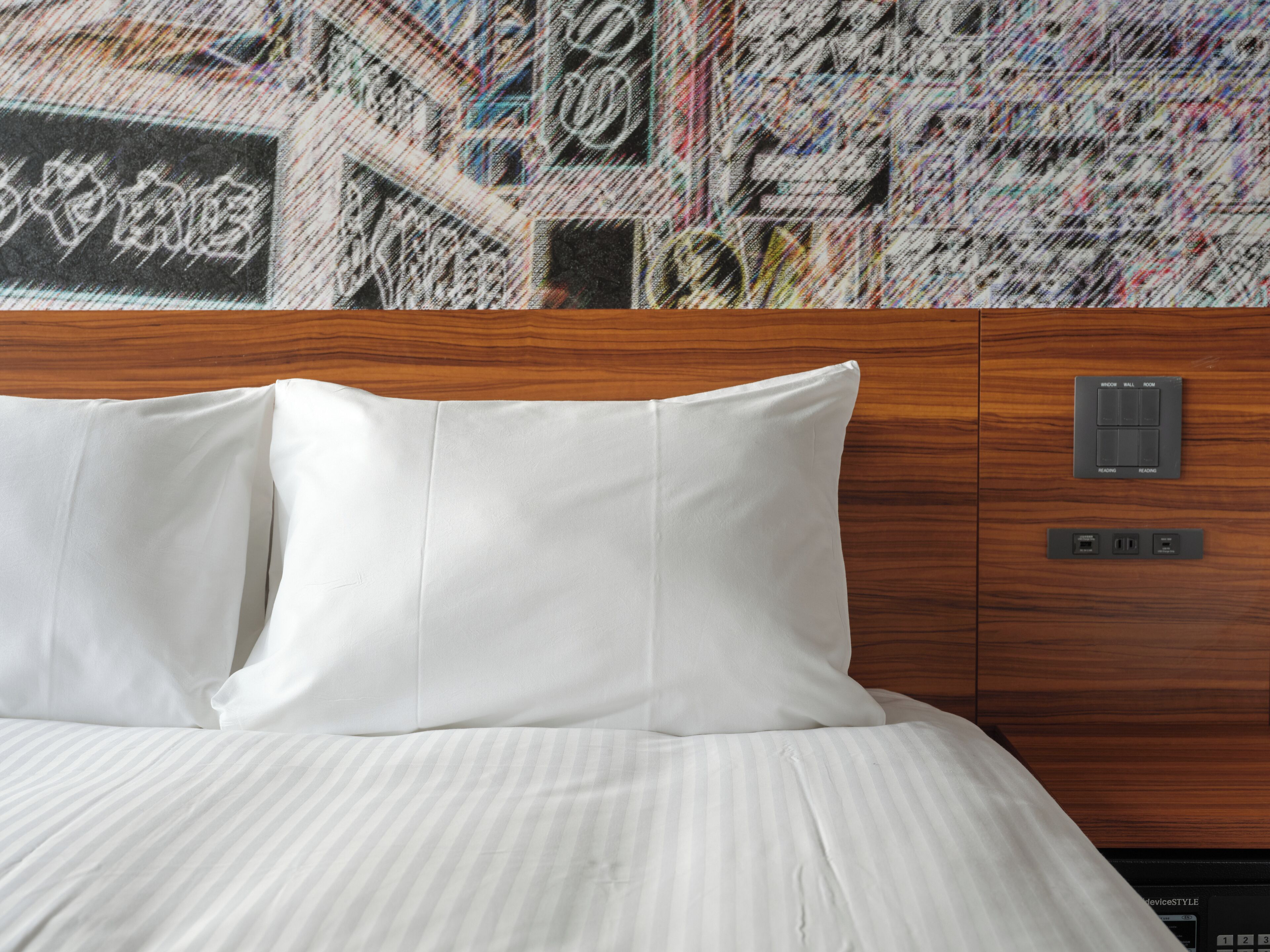 Ein Hotelzimmer mit einem hölzernen Kopfteil, weißen Kissen, einer gestreiften Bettdecke und einem Bedienfeld mit mehreren Knöpfen und einem Display.