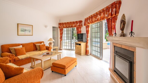 A living room with a sofa, ottoman, coffee table, TV, fireplace, and large windows with curtains. - Casa Oliveiras, villa with private pool & garden (Alvor)