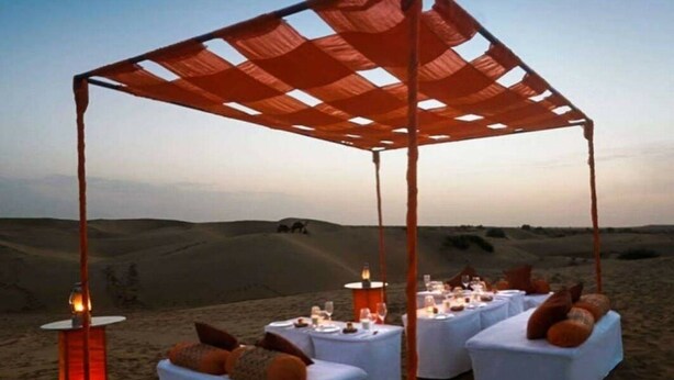 A desert tent set up with white cushioned seating and tables, a fire pit, and a view of the desert landscape. - Hotel Diamond Villa Safari Jaisalmer  (Jaisalmer)