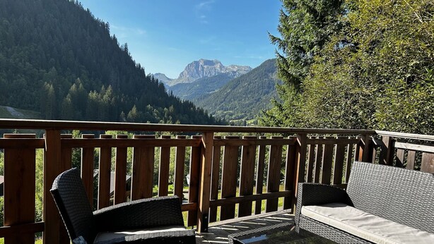 Een houten terras met rieten meubilair en een glazen tafel, met uitzicht op een bergachtig landschap.