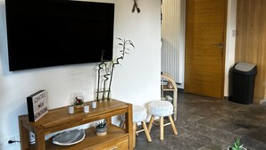 Een moderne woonkamer met een flatscreen televisie aan de muur, een houten console met lades, een klein krukje en een potplant.