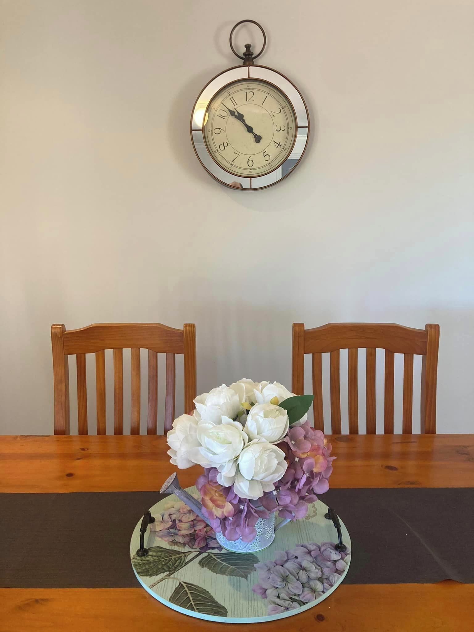 A dining table with a floral centerpiece, two wooden chairs, and a wall-mounted clock.