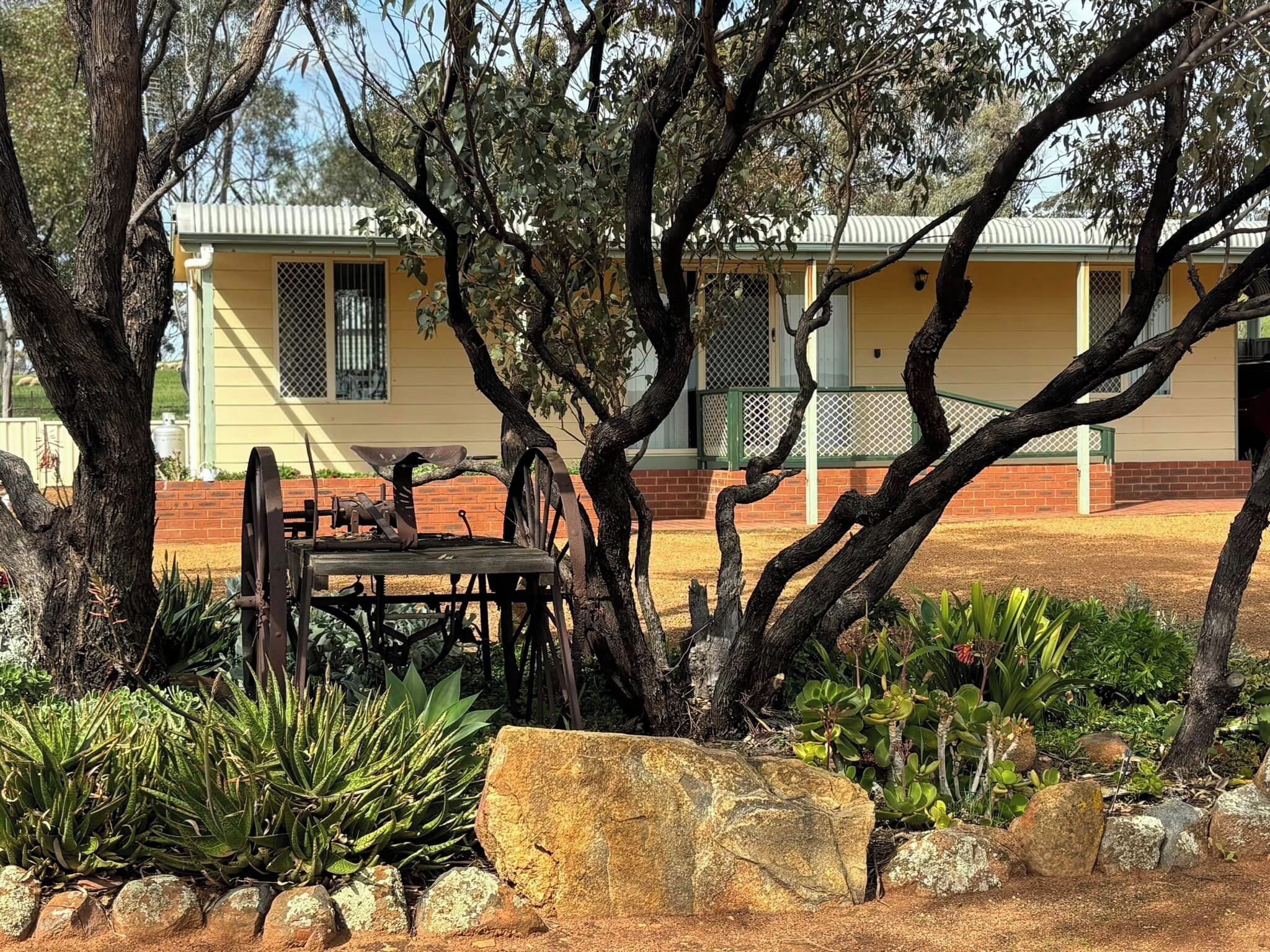 A rustic building with a yellow facade, surrounded by trees and a garden with rocks and plants.