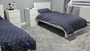 Une chambre avec deux lits simples, une table de chevet avec une lampe, une petite table avec une horloge et une fenêtre avec des rideaux.