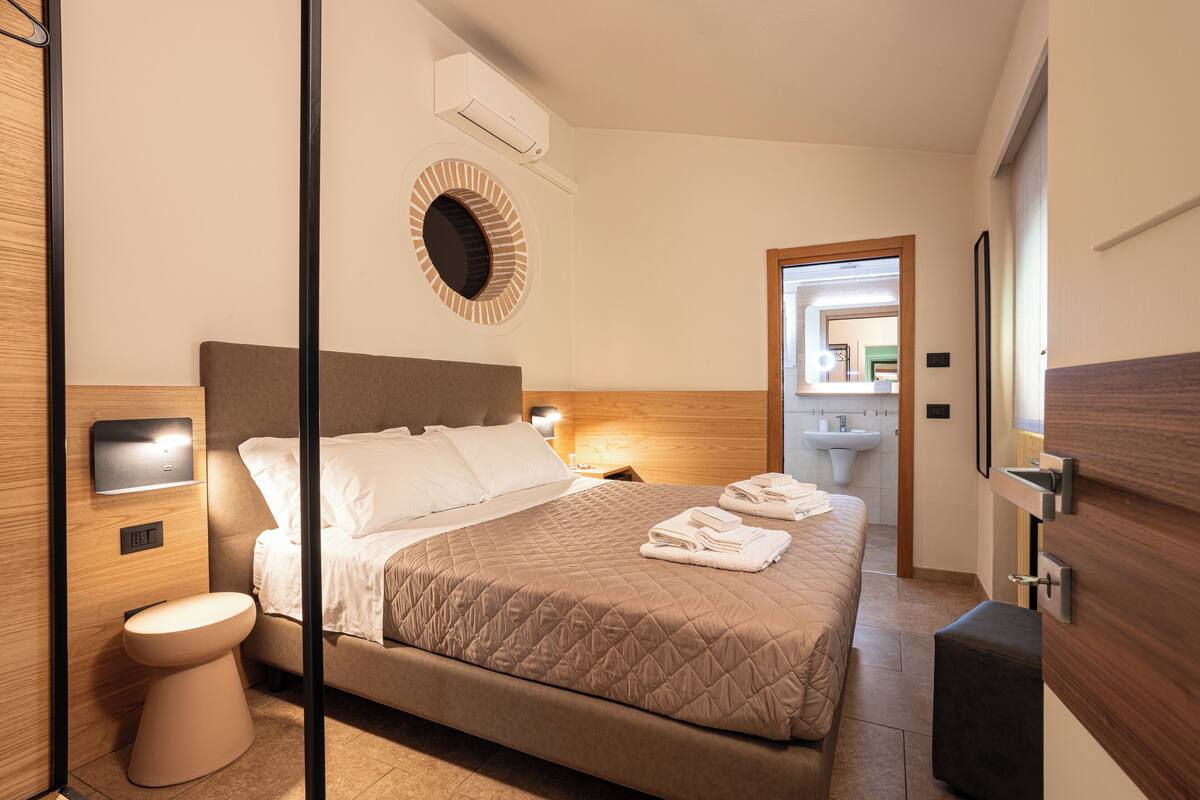 Une chambre d’hôtel moderne équipée d’un lit, d’une table de chevet, d’un climatiseur fixé au mur et d’un miroir rond placé au-dessus du lit.