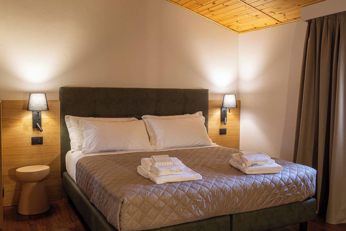 Un lit bien fait, avec une couette matelassée, deux oreillers et des serviettes pliées posées sur le lit. Deux lampes de chevet sont placées de chaque côté du lit.