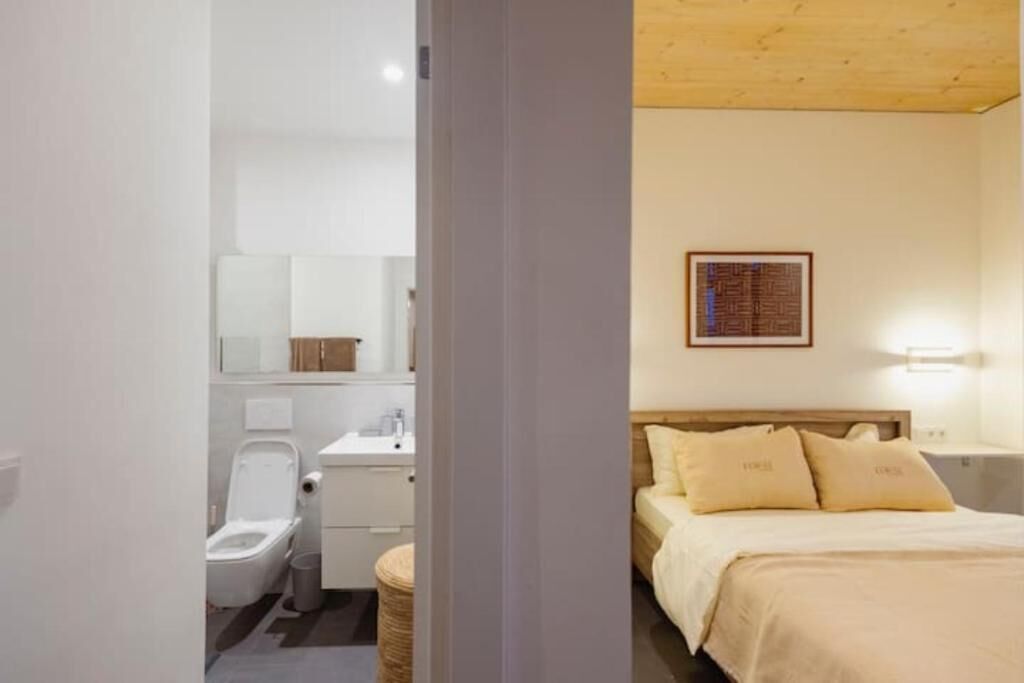 Un dormitorio con techo de madera, una cama con ropa de cama color beige y una mesita de noche con lámpara.