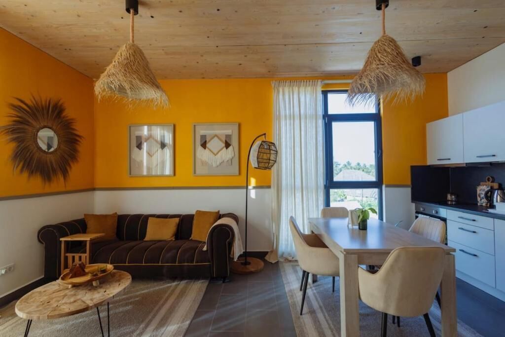 A modern living room with a sofa, dining table, and chairs. There is a window with curtains, a kitchen area, and a ceiling fan.