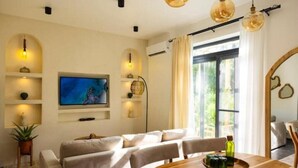 A modern living room with a wooden dining table, comfortable seating, a flat-screen TV, and a large window with curtains. - Nafsi Nomad (Paje)