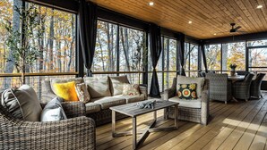 Un área de estar al aire libre techada con mobiliario de mimbre, una mesa y grandes ventanales con vistas a un bosque.