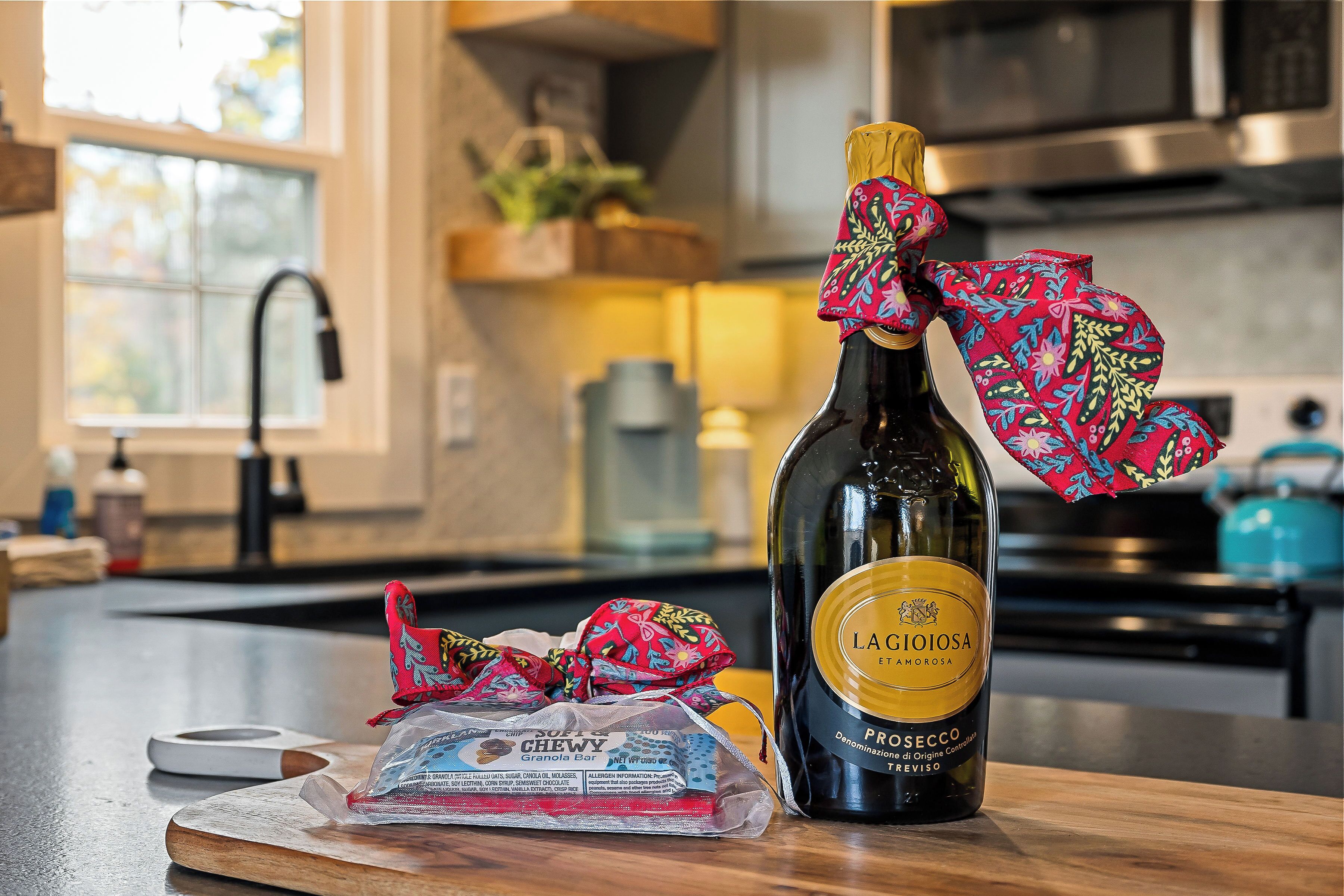 A bottle of LAGIOIOSA PROSECCO on a wooden surface with a colorful cloth tied around it, a granola bar, and a white object in a kitchen setting.