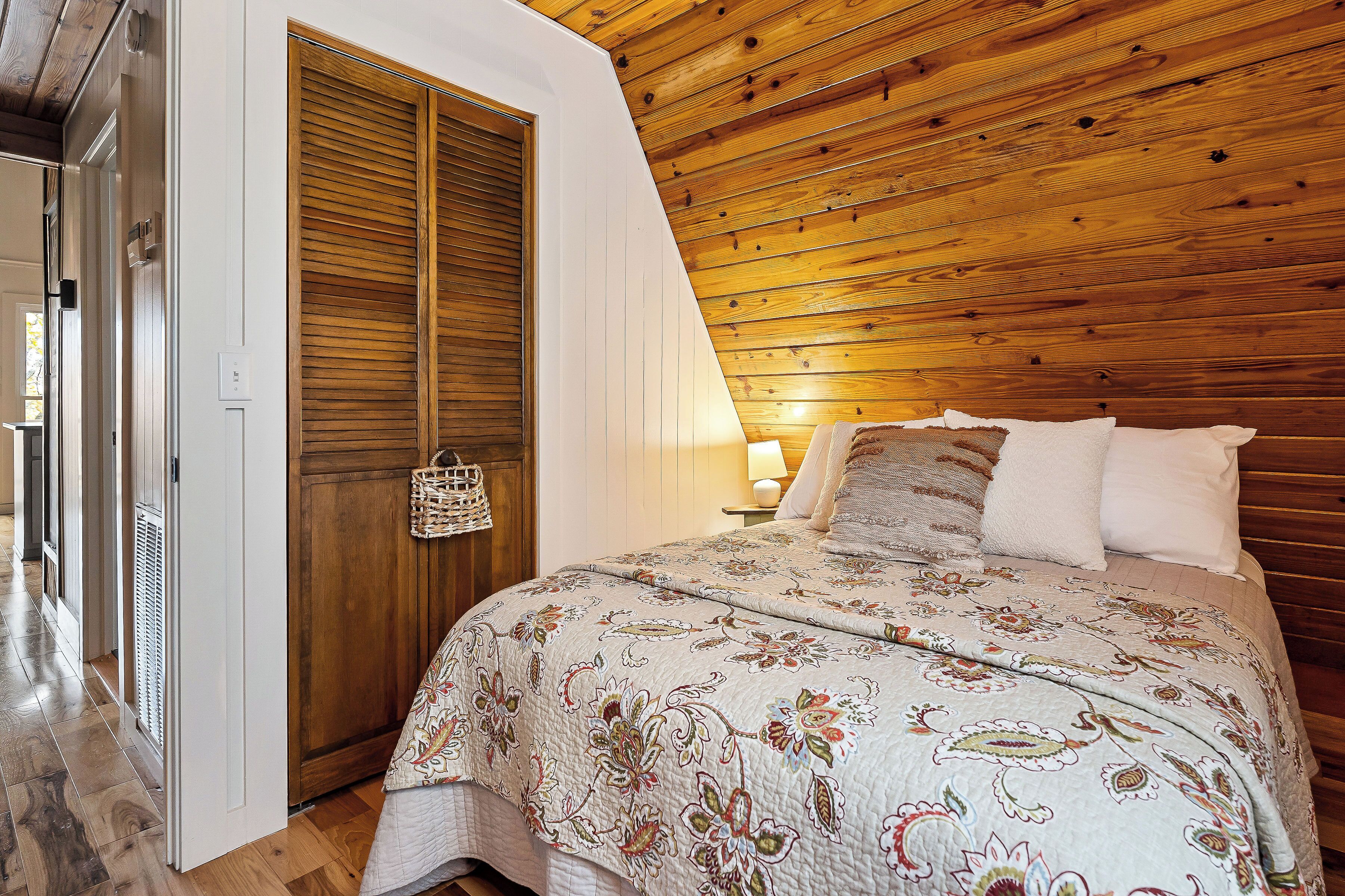 Una camera da letto con soffitto in legno, un letto con copripiumino floreale, una porta in legno e un comodino con una lampada.