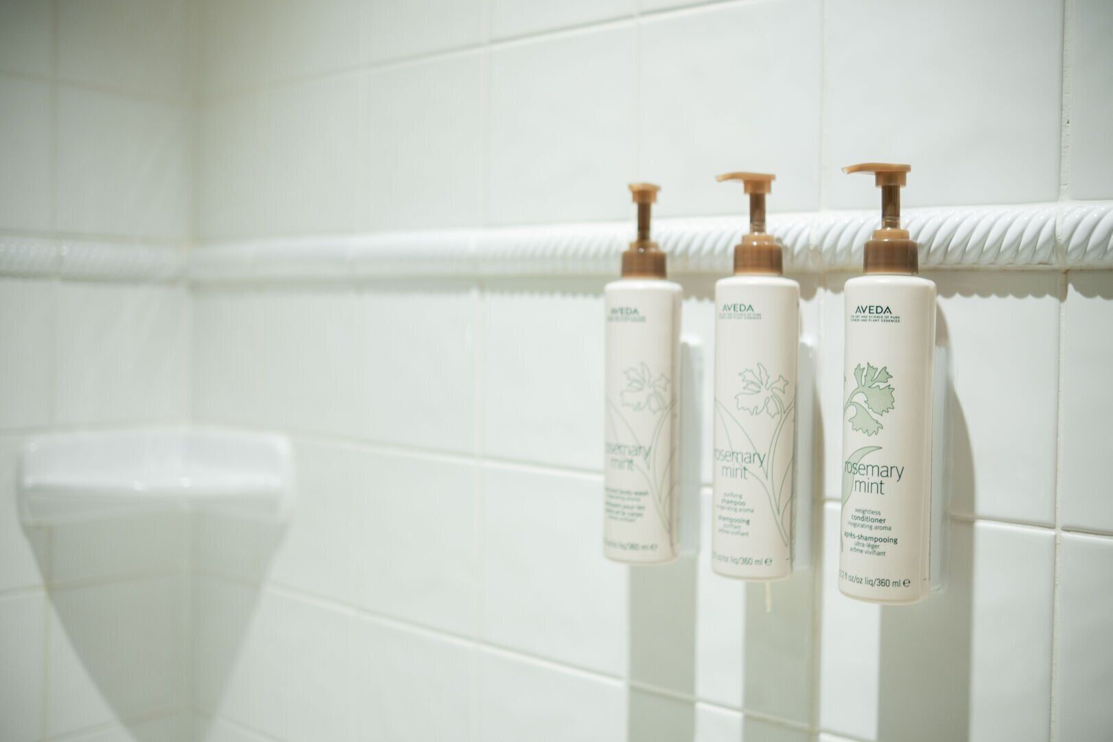 Three bottles of Aveda Rosemary Mint shampoo and conditioner on a white tiled wall.