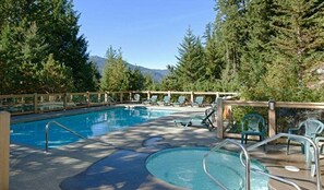 Area piscina con lettini e un pontile in legno, circondata da alberi e montagne.