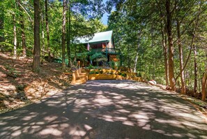 Uma cabana com telhado verde e varanda, cercada por árvores altas e um caminho pavimentado que a leva.