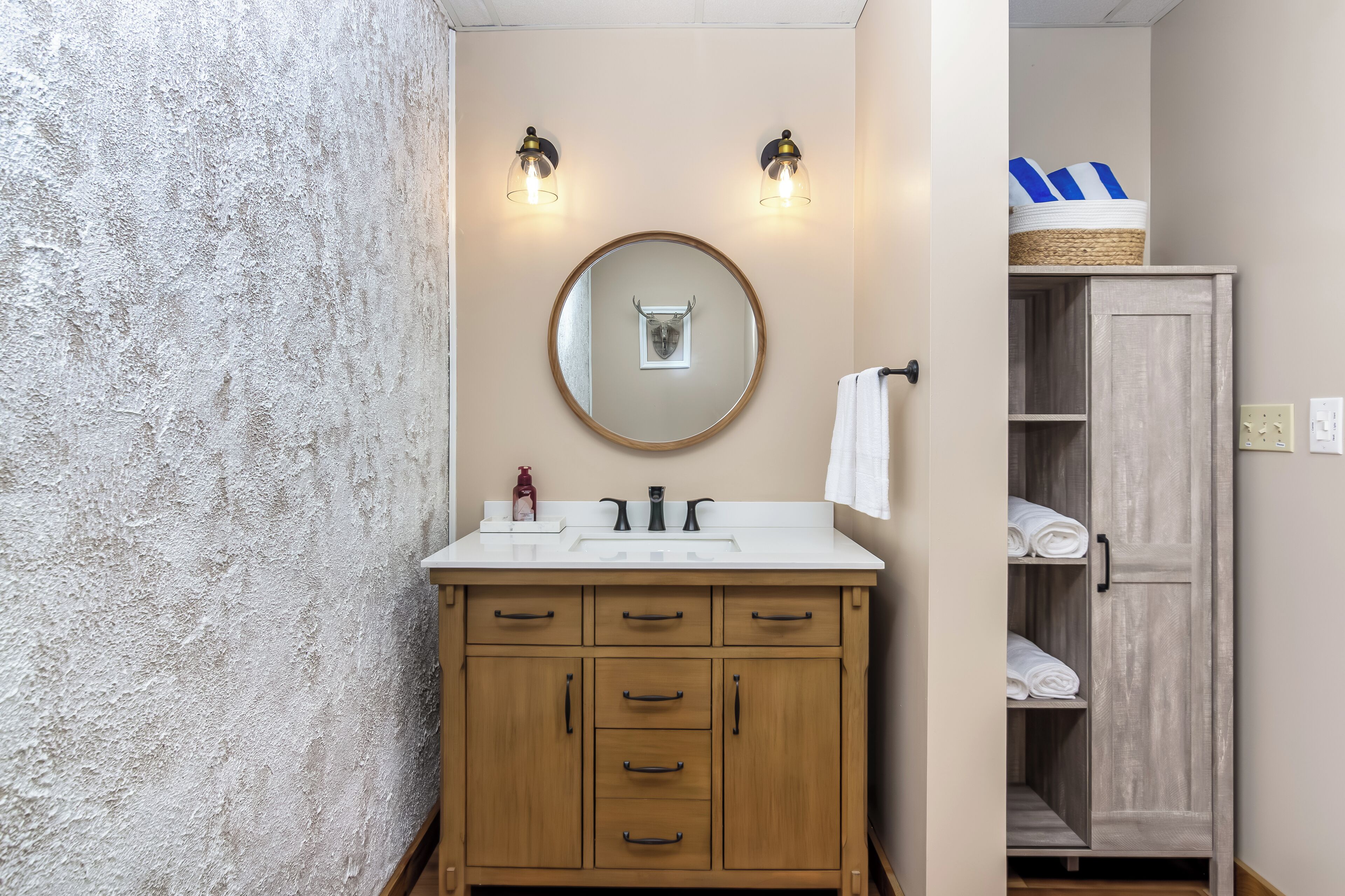 Une salle de bain avec un meuble-lavabo en bois, un miroir rond, deux appliques murales, un porte-serviettes et un meuble en bois contenant des serviettes.