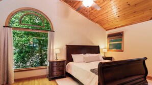 A bedroom with a wooden ceiling, a large window with a view of trees, a bed with white bedding, two bedside tables with lamps, and a painting on the wall. - New Listing! Mtn Cabin: Fire pit, Hot tub Views (Cherry Log)