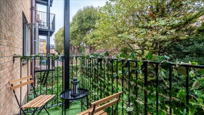 Un petit espace extérieur aménagé avec deux bancs en bois et une table ronde, entouré d’une clôture en métal et d’une végétation luxuriante.