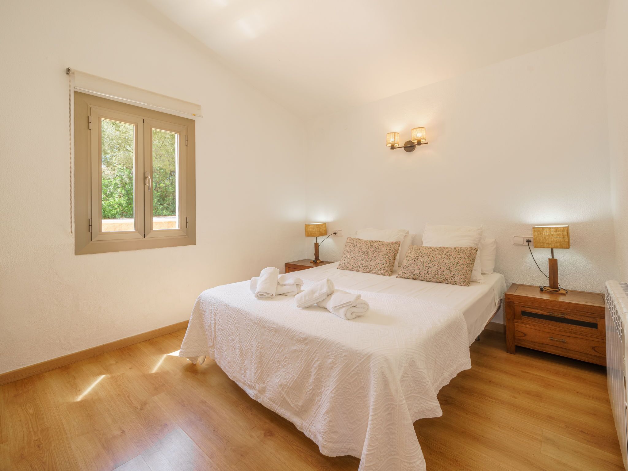 Un dormitorio con una cama grande, dos mesitas de noche con lámparas, una ventana que da a vistas de vegetación y suelo de madera.