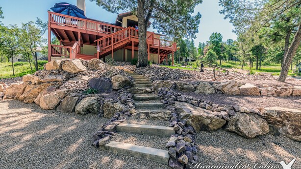 A cabin with a stone staircase leading to a wooden deck, surrounded by large rocks and trees. - Our Comfy Cabin | Cozy Retreat w/ Foosball, Wildlife Views & Family Fun (Ruidoso)