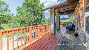 A wooden deck with a stone wall, outdoor furniture, and a view of trees. - Our Comfy Cabin | Cozy Retreat w/ Foosball, Wildlife Views & Family Fun (Ruidoso)