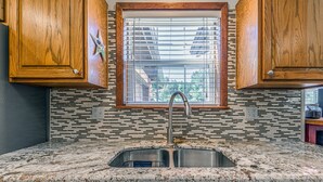 A kitchen with wooden cabinets, a window with blinds, a stainless steel sink, and a tiled backsplash. - Our Comfy Cabin | Cozy Retreat w/ Foosball, Wildlife Views & Family Fun (Ruidoso)