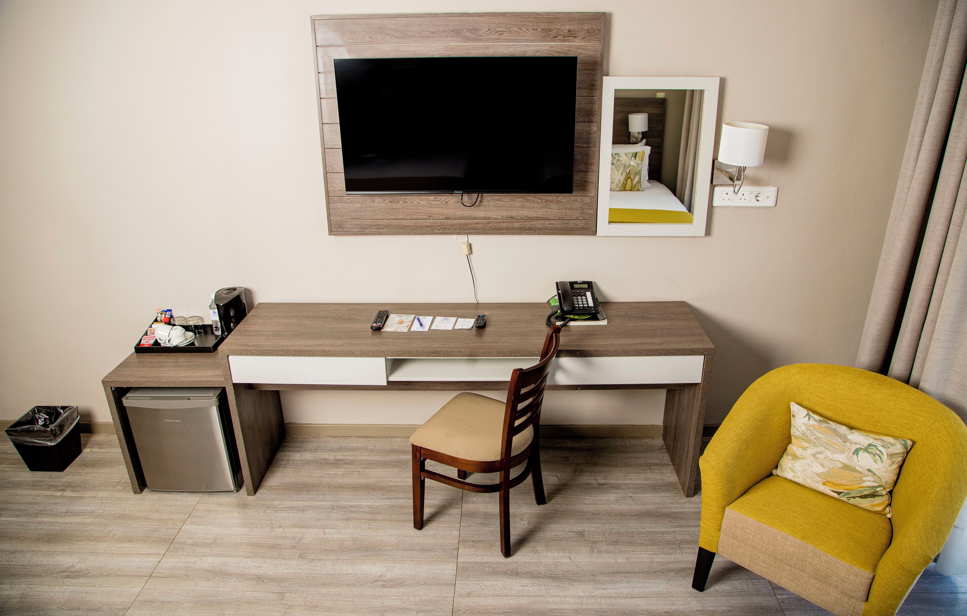 Camera d'albergo compatta con una scrivania in legno, una televisione, un piccolo frigorifero, una sedia e una poltrona gialla.