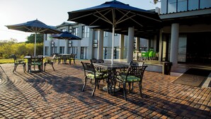 An outdoor patio area with brick flooring, tables, chairs, and umbrellas. - Two Rivers Hotel (Kasane)