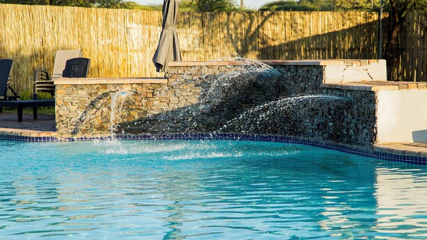A swimming pool with a stone wall and a wooden fence in the background. - Two Rivers Hotel (Kasane)