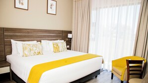 A hotel room with a large bed, a bedside table, a chair, and a window with curtains. - Two Rivers Hotel (Kasane)