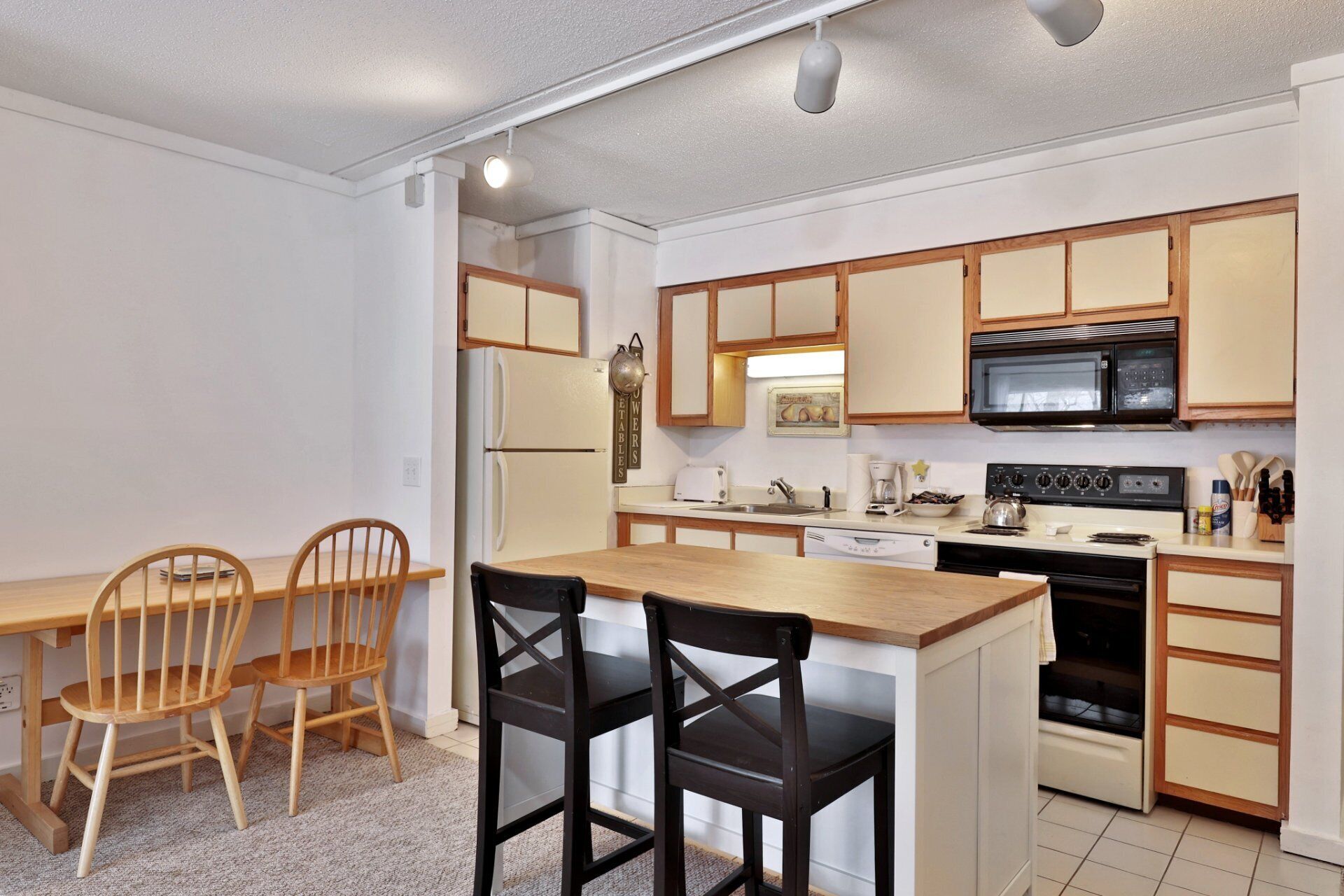 A compact kitchen with wooden cabinets, a refrigerator, a microwave, a stove, and a sink. There are two wooden chairs and a small island with a black chair.
