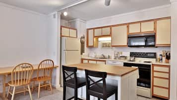 A compact kitchen with wooden cabinets, a refrigerator, a microwave, a stove, and a sink. There are two wooden chairs and a small island with a black chair.