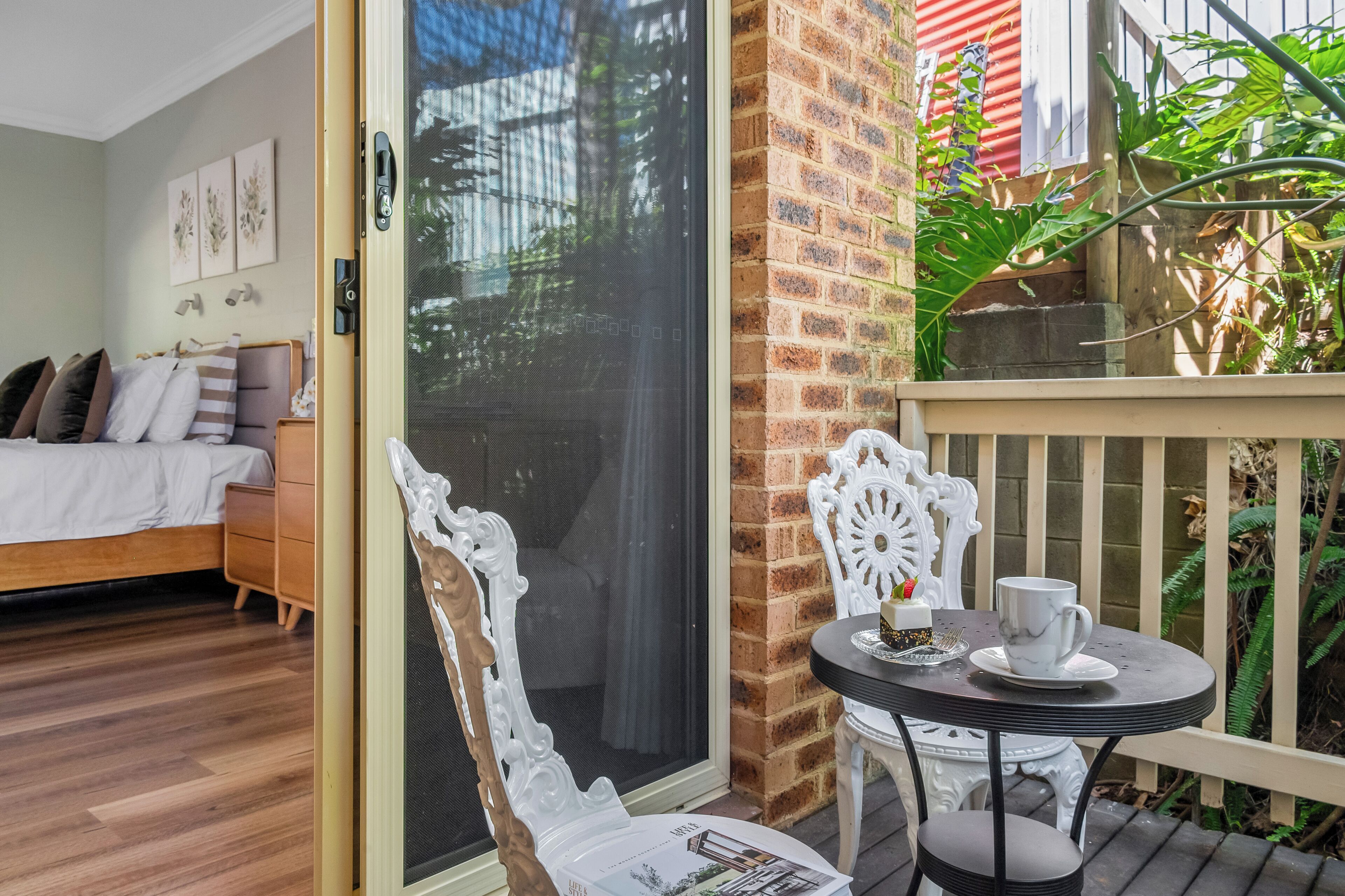 A bedroom with a bed, a wooden dresser, and a sliding glass door leading to an outdoor area with a table, chairs, and a potted plant.