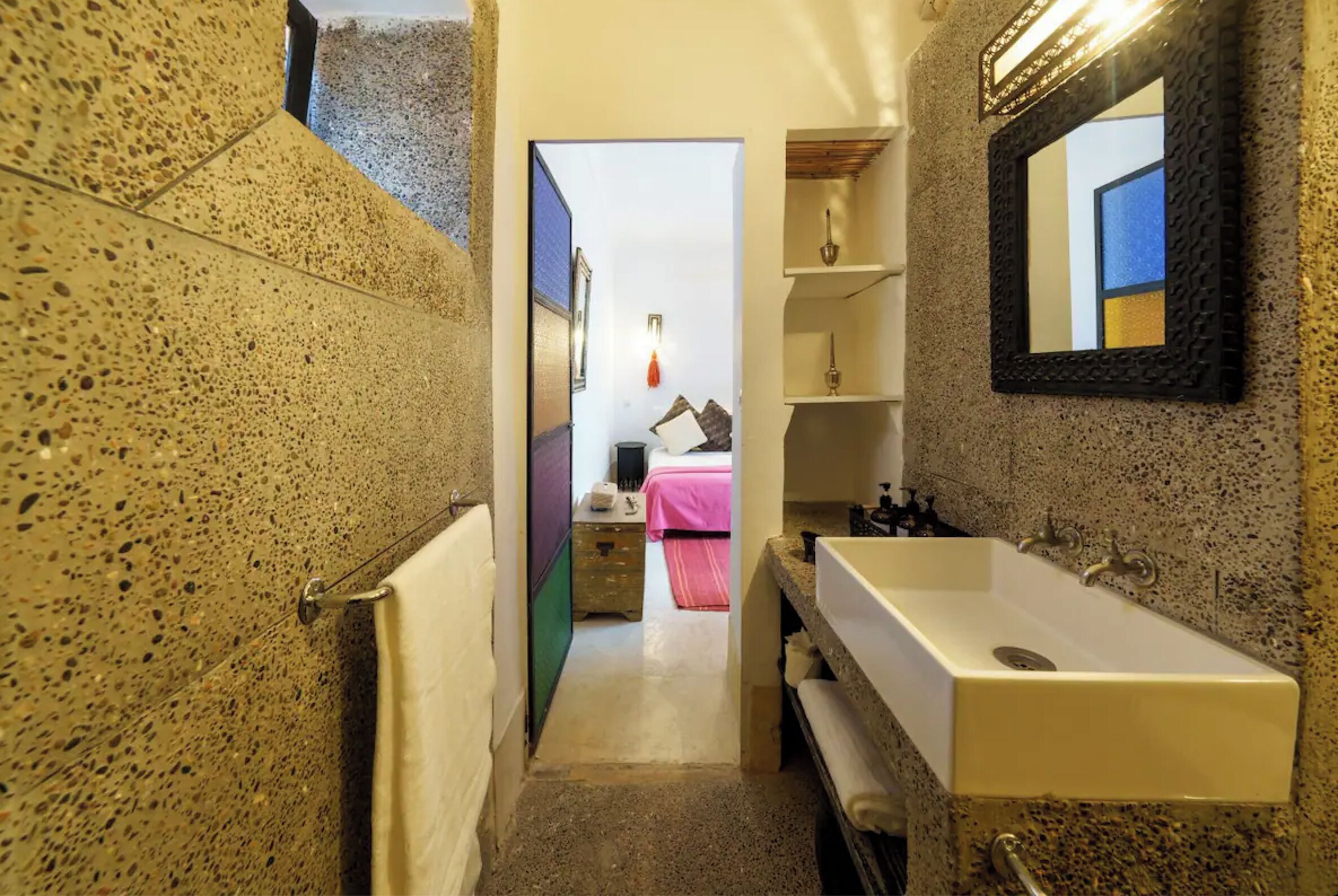 Un couloir étroit mène à une chambre à coucher qui contient un lit, une table de chevet et une commode. On y trouve également une salle de bain avec un lavabo, un miroir et des étagères.