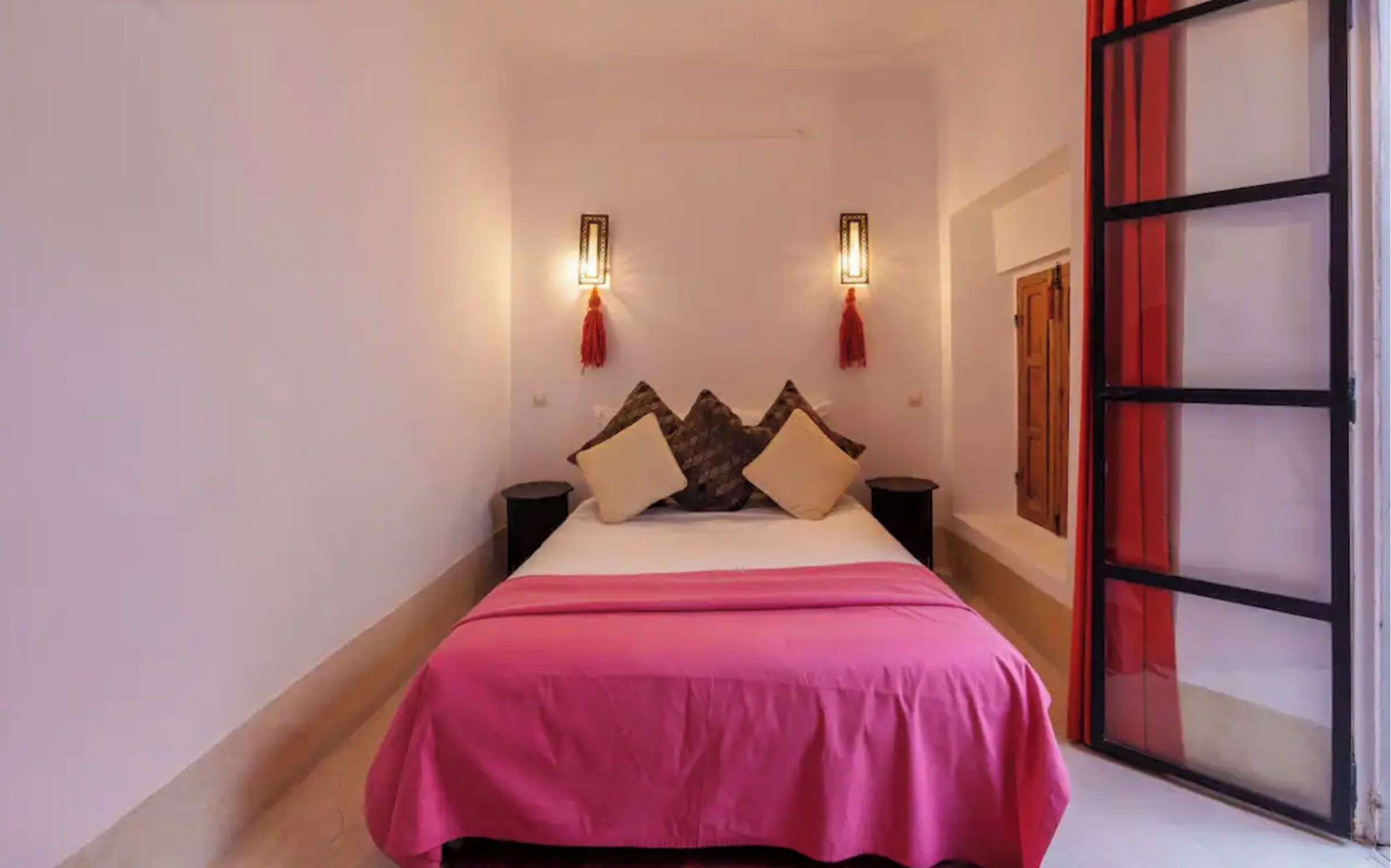 A narrow hotel room with a bed, two wall-mounted lights, and a red curtain.