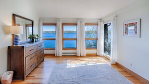 Ein Zimmer mit einem Holzkoffer, einem Spiegel, einer Lampe, einem Teppich und großen Fenstern mit Blick auf einen See und Bäume.