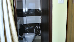 A narrow bathroom with a toilet, a blue bucket, and a trash bin. - MM Hotel and Service Apartment (Kolkata)