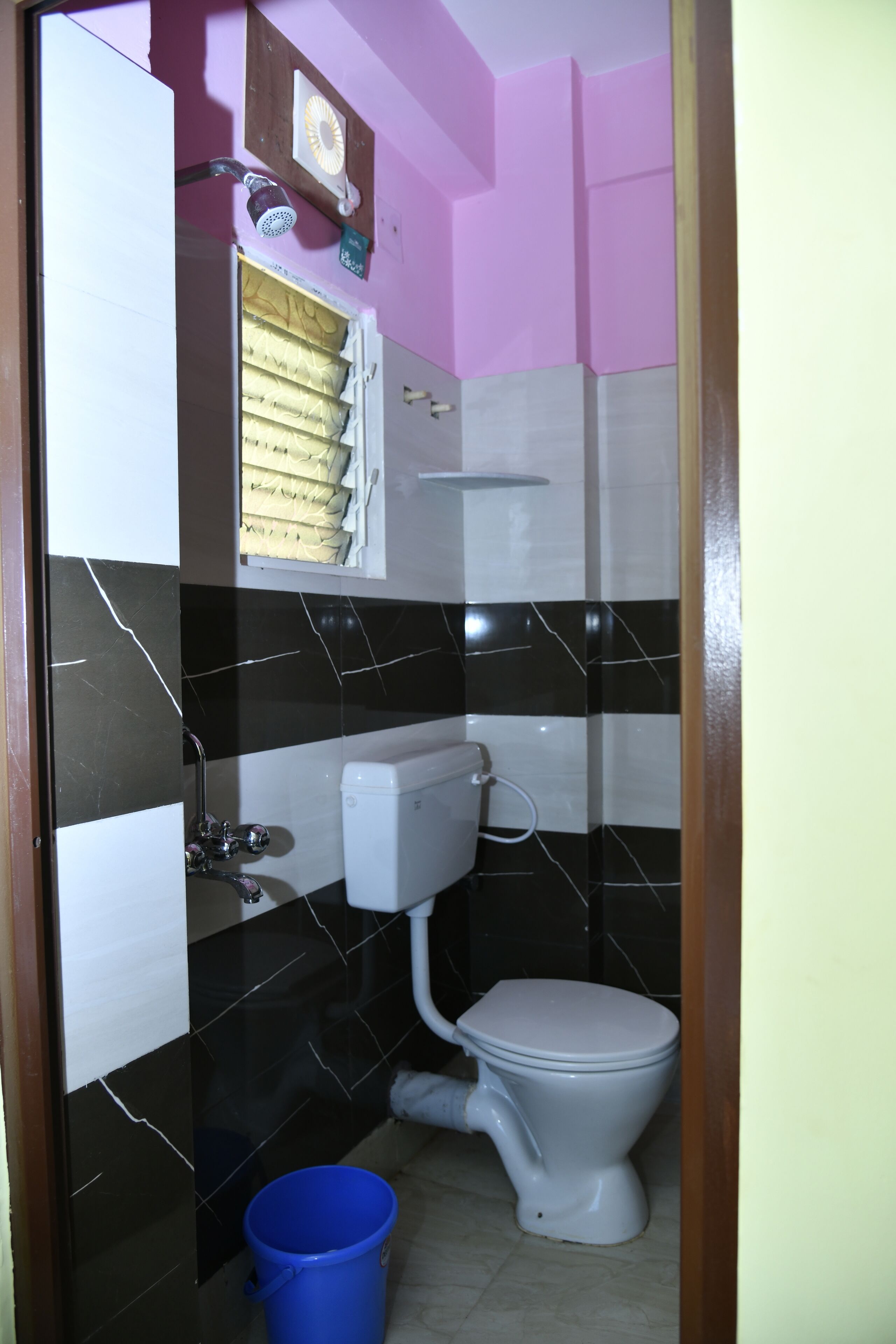 Une salle de bain avec des toilettes, un seau bleu et une fenêtre avec des stores.