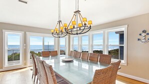 Un'area pranzo con tavolo in vetro e sedie in vimini, ampie finestre vista oceano e un lampadario.