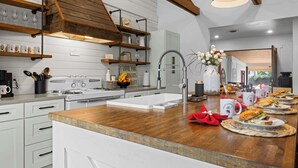 A modern kitchen with white cabinetry, wooden beams, a stainless steel sink, and a breakfast setup with plates, mugs, and a vase of flowers. - Kilted Cowboy - Cowboy Pool, Hot Tub & Fire Pit (Fredericksburg)