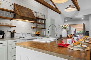 A modern kitchen with white cabinetry, wooden beams, a stainless steel sink, and a breakfast setup with plates, mugs, and a vase of flowers.