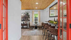 A room with a wooden ceiling, a red door, a sofa, a coffee table, and a bar area with stools. - Kilted Cowboy - Cowboy Pool, Hot Tub & Fire Pit (Fredericksburg)