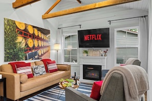 A living room with a sofa, a coffee table with a bottle and fruit, a fireplace, and a TV displaying 'NETFLIX'. - Kilted Cowboy - Cowboy Pool, Hot Tub & Fire Pit (Fredericksburg)