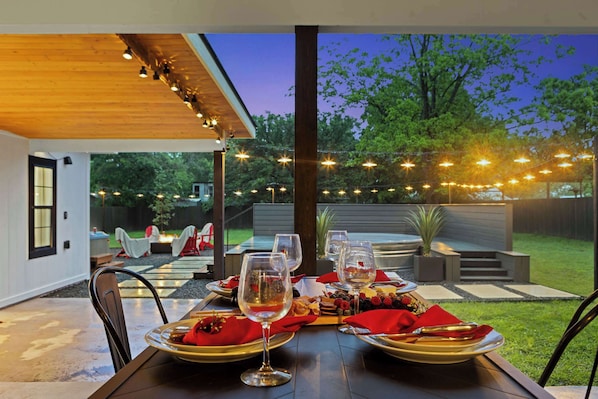 An outdoor dining area with a table set for two, surrounded by string lights and a view of the backyard.