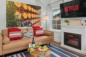 A living room with a beige sofa, red and white pillows, a wine bottle, glasses, a fruit platter, a fireplace, a Netflix TV, and a large wine barrel painting. - Kilted Cowboy - Cowboy Pool, Hot Tub & Fire Pit (Fredericksburg)