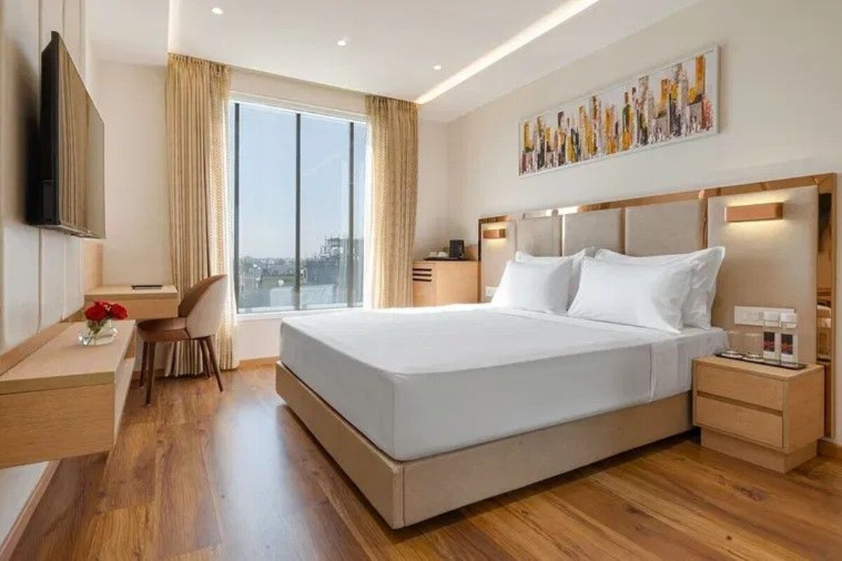 A modern hotel room with a large bed, a desk, a chair, a TV, and a city view.